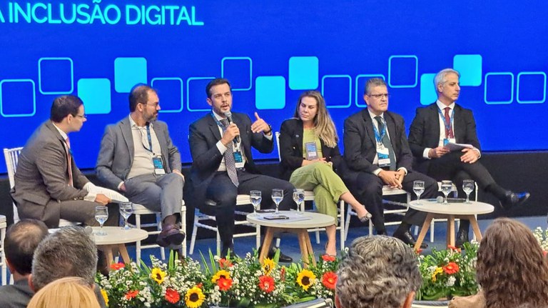 Foto de um painel de discussão com cinco palestrantes sentados no palco. Ao centro, o conselheiro da Anatel, Edson Holanda, fala ao microfone e gesticula com a mão direita. Ele participa do painel "Cabos Submarinos e Data Centers: Políticas Nacionais para Inclusão Digital", promovido pelo Ministério das Comunicações. O painel também inclui outros três homens e uma mulher. O fundo é uma tela azul brilhante que exibe a frase "INCLUSÃO DIGITAL". Em primeiro plano, há arranjos florais coloridos e a parte de trás da cabeça da plateia.
