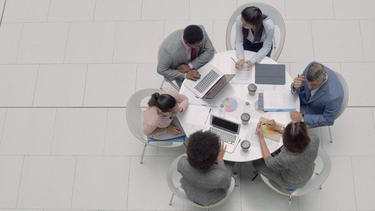 À direita da imagem pessoas em uma mesa redonda com papéis e laptops.