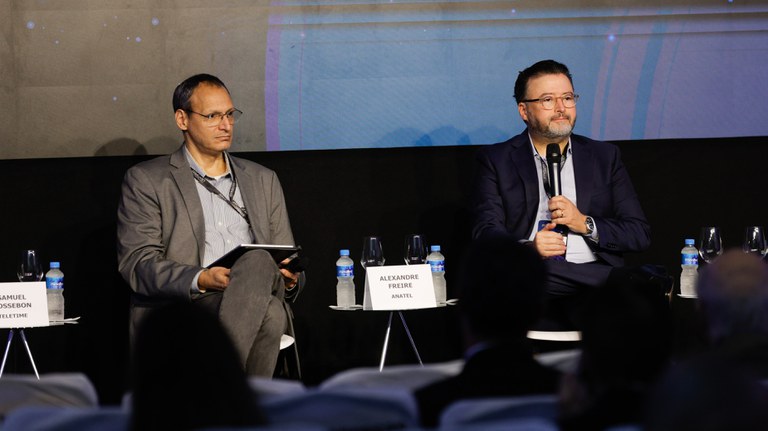 Dois homens em um seminário. O da direita, o conselheiro Alexandre Freire, está vestido com um terno azul-marinho. Ele gesticula com a mão direita enquanto fala, sentado em uma cadeira branca. O homem à esquerda veste um blazer cinza e calças jeans escuras, sentado com as pernas cruzadas. Ambos estão em frente a um painel escuro com pontos de luz simulando estrelas. Há pequenas mesas laterais de metal entre eles com garrafas de água e taças.