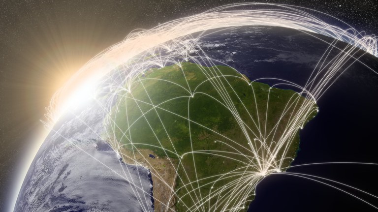 Uma imagem de satélite da Terra, focada na América do Sul, com linhas brancas brilhantes que representam conexões de rede ou rotas de voo irradiando-se sobre o continente e ao redor do globo. O sol está nascendo ou se pondo no horizonte, criando um brilho intenso no lado esquerdo da imagem.