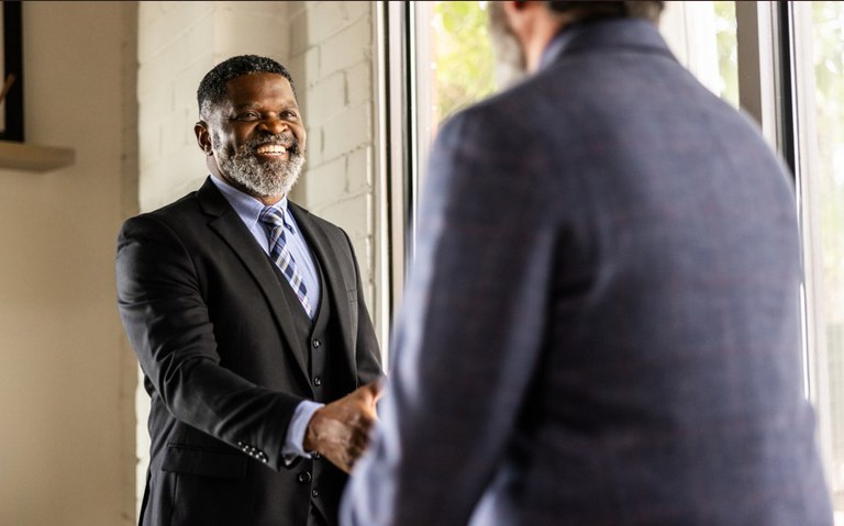 Um homem negro de meia-idade, com barba grisalha e um sorriso largo, está de pé e aperta a mão de um homem branco mais velho, que está de costas para a câmera. Ambos usam ternos formais. A cena se passa em um escritório com uma parede de tijolos expostos e uma grande janela. O foco está no homem negro sorridente, enquanto o outro homem está em desfoque.