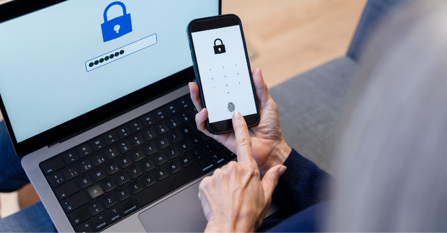 Pessoa segura um smartphone exibindo tela de bloqueio com ícone de cadeado e teclado numérico, enquanto toca no ícone de impressão digital. Ao fundo, um notebook mostra uma tela com cadeado azul e campo de senha preenchido com pontos. A cena representa autenticação segura.