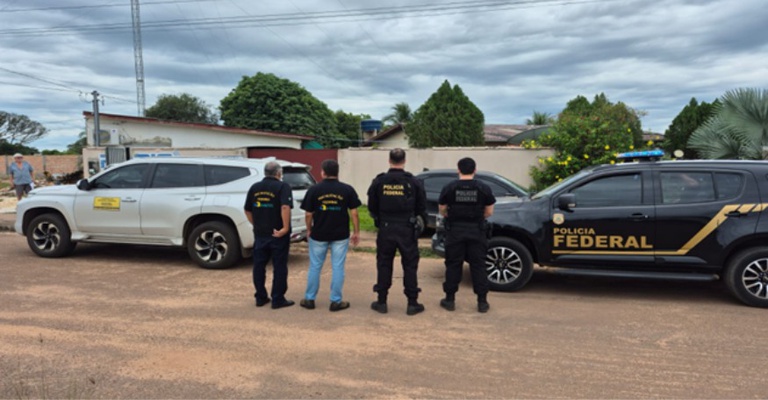Agentes da Anatel e da Polícia Federal aparecem de costas, lado a lado, em uma via urbana, durante operação de fiscalização em Terra Nova do Norte (MT). Duas viaturas oficiais estão estacionadas à frente dos agentes, representando ação conjunta de combate a emissora de rádio clandestina.