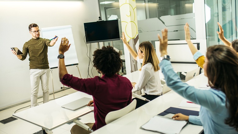 Imagem mostra estudantes levantando os braços para fazer perguntas ao professor em uma sala de aula