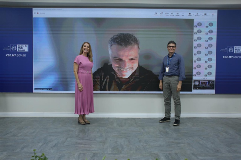 Imagem mostra uma mulher e um homem no palco, com um telão mostrando uma terceira pessoa