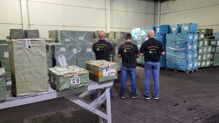 rês homens com camisetas pretas da "FISCALIZAÇÃO FEDERAL" e logotipo da Anatel observam um armazém repleto de pilhas de caixas grandes embrulhadas em plástico verde e azul, organizadas em paletes. No primeiro plano, caixas menores com etiquetas brancas e números como "89" e "68" estão sobre uma mesa de metal. O chão é de paralelepípedos.