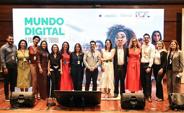 Fotografia de um grupo de pessoas em um palco, posando lado a lado para uma foto oficial. Ao todo, há cerca de 15 adultos, homens e mulheres, vestidos de forma variada — desde roupas sociais formais até trajes mais leves e coloridos. Alguns usam crachás pendurados no pescoço, indicando participação em evento institucional. Ao fundo, há um grande telão com a inscrição “MUNDO DIGITAL” em destaque, seguida de um subtítulo parcialmente visível. Também aparecem logotipos institucionais, incluindo o da Anatel e do Governo Federal. No telão, há ainda a ilustração de uma personagem feminina estilizada, com traços modernos, associada ao tema digital.