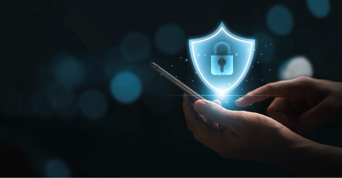 Uma mão segurando um smartphone com um escudo digital luminoso e um cadeado, simbolizando segurança e proteção de dados.