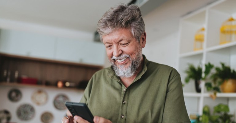 A imagem mostra um homem de meia-idade sorridente, com barba e cabelos grisalhos, usando uma camisa verde. Ele está segurando um celular e olhando para a tela com expressão de alegria ou satisfação.