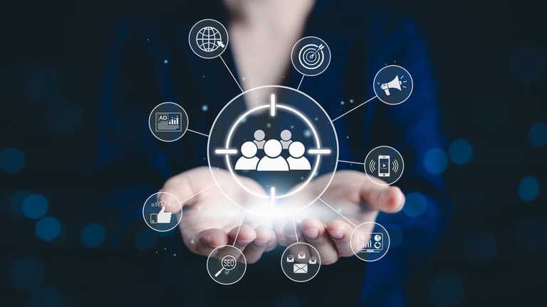 Uma pessoa veste um blazer azul escuro e segura as mãos abertas, de onde parece emanar um brilho central. Sobre as mãos, flutua um gráfico digital composto por um alvo central contendo o ícone de três pessoas (representando o público-alvo). Ao redor desse alvo, estão dispostos diversos ícones de marketing conectados por linhas finas