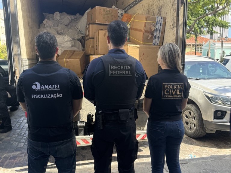 Três agentes de fiscalização brasileiros, um homem da ANATEL, um homem da RECEITA FEDERAL e uma mulher da POLÍCIA CIVIL PERNAMBUCO, estão de costas, observando o interior de um caminhão baú cheio de caixas de papelão empilhadas. A cena ocorre ao ar livre, com uma caminhonete prateada estacionada ao lado do caminhão.