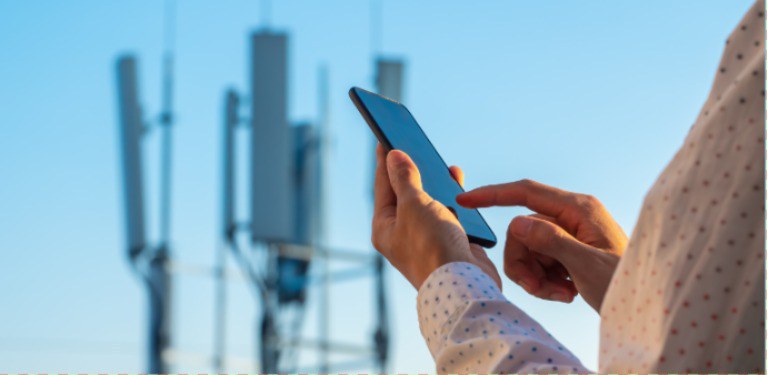 Imagem destaca as mãos de uma pessoa utilizando celular com uma torre e antenas de telecomunicações ao fundo.