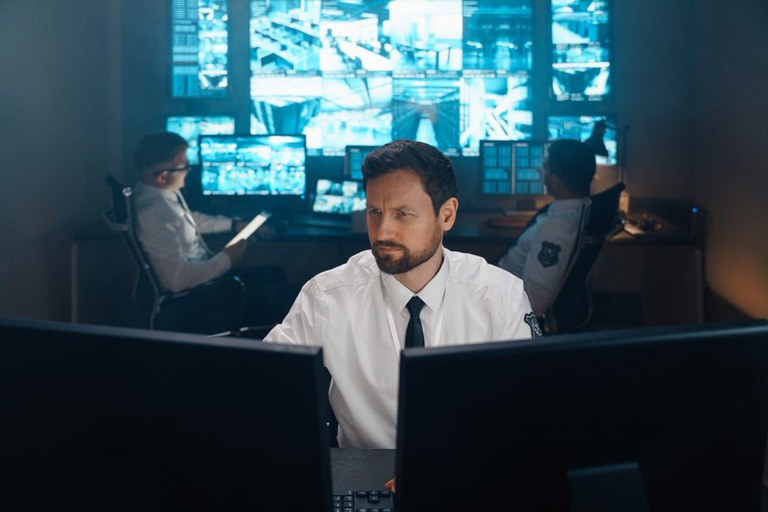 Três homens em uma central de monitoramento com telas de vigilância