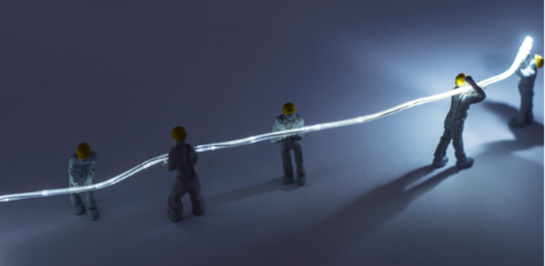 A imagem apresenta uma cena conceitual na qual pequenas figuras de trabalhadores, usando capacetes amarelos e roupas de proteção, manipulam um cabo de fibra óptica iluminado, simbolizando o trabalho de pequenos provedores de telecomunicações, que atuam na manutenção e expansão das redes de internet e promoção da conectividade.