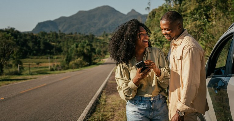 Na imagem, casal sorri ao lado do carro em uma estrada cercada por vegetação, e montanhas são visíveis no horizonte. Ambos sorriem enquanto a mulher segura um celular e o homem olha para o aparelho.