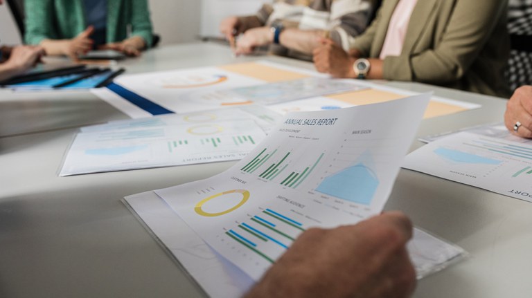 A imagem captura uma reunião de negócios com foco na análise de relatórios de vendas. Em primeiro plano, as mãos de uma pessoa seguram um documento intitulado "ANNUAL SALES REPORT", que apresenta vários gráficos coloridos, incluindo gráficos de barras, um gráfico de pizza amarelo e um gráfico de área azul. Outros documentos semelhantes com gráficos e tabelas estão espalhados pela mesa de conferência cinza, juntamente com tablets e canetas. Várias pessoas estão sentadas ao redor da mesa, vestindo roupas casuais de negócios, algumas gesticulando e interagindo com os materiais, enquanto outras observam. A cena é bem iluminada, com luz natural vinda da esquerda.