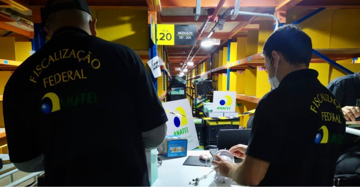Uma fotografia colorida captura dois agentes durante uma operação de fiscalização e autoridades federais em um grande armazém de distribuição