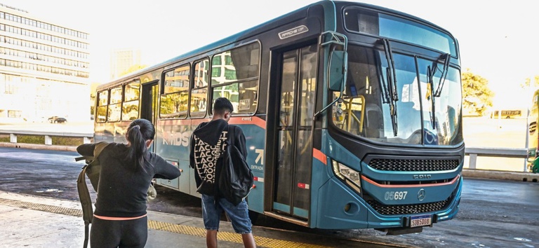 IMAGEM DE ONIBUS NA RODOVIARIA COM DUAS PESSOAS ESPERANDO EM PÉ