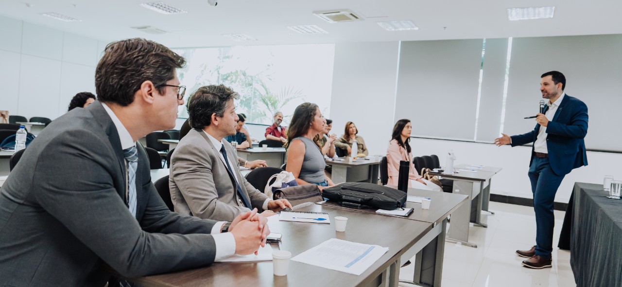 IMAGEM COM PESSOAS SENTADAS NA SALA COM UM HOMEM EM PÉ COM MICROFONE