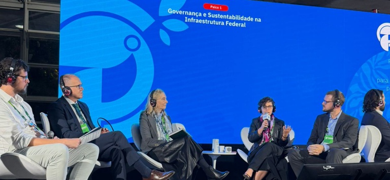 A imagem mostra um painel de discussão com cinco participantes sentados em palco, usando fones de ouvido. O telão ao fundo destaca o tema "Governança e Sustentabilidade na Infraestrutura Federal" durante o evento.