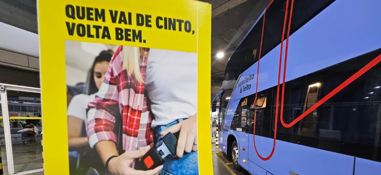 Foto mostrando folheto da campanha vou de cinto com um ônibus interestadual ao fundo