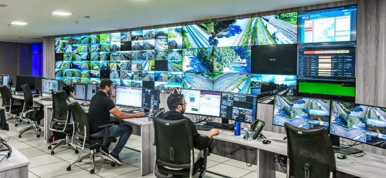 IMAGEM COM TRES PESSOAS SENTADAS EM CADEIRAS DE FRENTE PARA O COMPUTADOR E COM IMAGENS DAS RODOVIAS E FERROVIAS