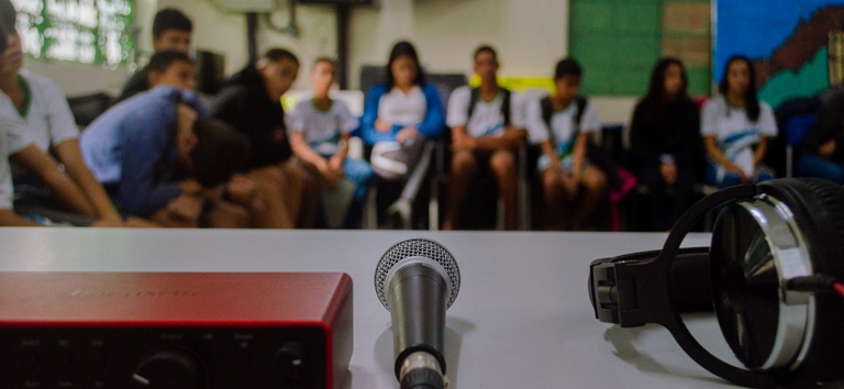 IMAGEM DE UM MICROFONE, UM HEADPHONE COM PESSOAS NO FUNDO DA FOTO