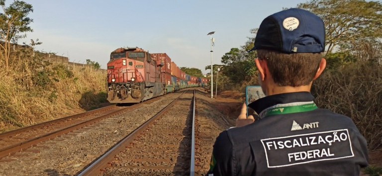Foto mostrando fiscal da ANTT, uniformizado, trabalhando em uma ferrovia com trem ao fundo