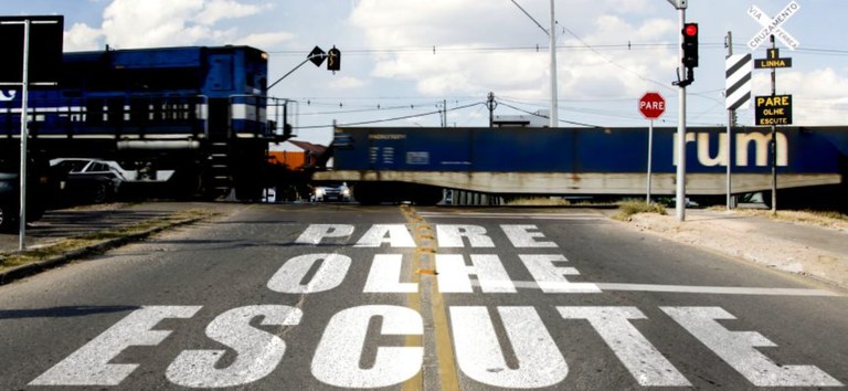 Foto mostrando trem passando em passagem de nível, cruzamento com carros e pedestres