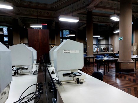 A imagem exibe o interior de uma biblioteca. Em primeiro plano, há uma bancada com equipamentos brancos que são máquinas leitores de rolos de microfilmes ou microfichas, voltados para o usuário. Os equipamentos estão alinhados, com fios visíveis conectando-os à mesa.Ao fundo, é possível ver mesas de estudo com cadeiras dispersas, em um ambiente amplo e iluminado. As paredes possuem detalhes arquitetônicos, com colunas em tons marrons e um teto decorado. As janelas, que permitem a entrada de luz natural, estão posicionadas em diferentes pontos do espaço, oferecendo uma sensação de amplitude. As janelas são altas, com moldura em cor verde e em boa parte constituída de vidro. O piso é de madeira, com um padrão que confere elegância ao ambiente. A atmosfera é tranquila, ideal para leitura e estudo.