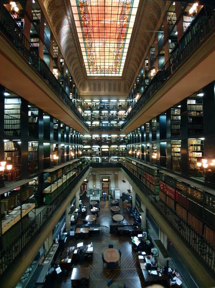 A imagem apresenta uma biblioteca grande e majestosa, o armazém de publicações seriadas da Biblioteca Nacional. com uma arquitetura rica e detalhada. O ambiente é centralizado em um espaço amplo, onde se pode ver várias estantes de periódicos, ou seja, jornais e revistas, que vão do chão ao teto, organizadas em prateleiras negras. Na parte superior, destaca-se um vitral colorido (Claraboia) que ilumina o espaço com uma luz suave e vibrante, criando um contraste com o escuro das estantes. As estantes são repletas de periódicos de diferentes tamanhos e cores. Abaixo, há mesas de estudo dispostas em grupos, com pessoas trabalhando em seus laptops ou lendo. As mesas, de onde foi fotografado o armazém de periódicos trata-se do salão de consulta de Manuscritos. O piso é de madeira, com um padrão que adiciona um toque de elegância ao ambiente. As paredes são adornadas com iluminação suave, e há um ar de tranquilidade e concentração. O teto é alto e decorado, conferindo uma sensação de grandiosidade ao espaço.