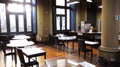 A imagem retrata um ambiente interno com mesas e cadeiras dispostas de maneira organizada. Há grandes janelas que permitem a entrada de luz natural, criando uma atmosfera clara e acolhedora. O piso é de madeira, e as mesas parecem ser de material branco, com algumas superfícies transparentes, possivelmente de vidro. O espaço parece tranquilo e é ideal para leitura ou estudo, com paredes em tons neutros e detalhes em azul nas janelas. A disposição dos móveis sugere um ambiente funcional e acessível para as atividades.
