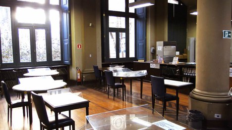 A imagem retrata um ambiente interno com mesas e cadeiras dispostas de maneira organizada. Há grandes janelas que permitem a entrada de luz natural, criando uma atmosfera clara e acolhedora. O piso é de madeira, e as mesas parecem ser de material branco, com algumas superfícies transparentes, possivelmente de vidro. O espaço parece tranquilo e é ideal para leitura ou estudo, com paredes em tons neutros e detalhes em azul nas janelas. A disposição dos móveis sugere um ambiente funcional e acessível para as atividades.