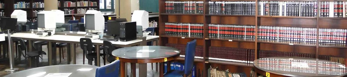 A imagem captura parte do salão de leitura de Publicações Seriadas e Referência da Biblioteca Nacional. No primeiro plano, há mesas redondas de madeira com tampo de vidro, acompanhadas de cadeiras azuis. No segundo plano, estantes de madeira exibem periódicos encadernados com lombadas em vermelho e preto ou marrom. À esquerda, uma fileira de máquinas leitoras de microfilmes e computadores. Mais ao fundo, no terceiro plano, estantes de madeira abrigam obras de referência.