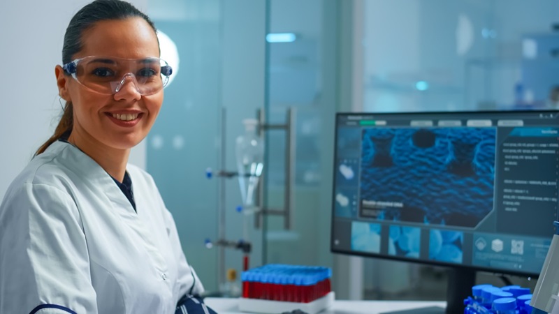 Cientista de jaleco e óculos de proteção sorri em um laboratório moderno. Ao seu lado, há um computador exibindo dados e imagens de análises clínicas, com tubos de ensaio sobre a bancada.