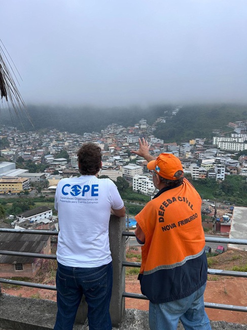 Foto 3- Pesquisador do Projeto COPE recebe explicação do agente da defesa civil sobre as áreas mais atingidas pelo desastre de 2011, em Nova Friburgo -RJ. (Foto:Victor Marchezini-Projeto COPE Cemaden).#PraTodosVerem:A direita, um homem de costas com o colete laranja da defesa civil, aponta para o horizonte, enquanto outro homem a esquerda com blusa do Projeto COPE observa a cidade. Ao fundo, as casas e prédios estão em uma região de mata com muita neblina.