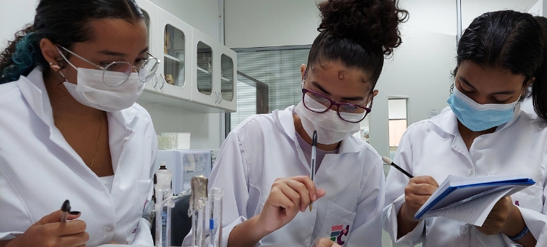 Estudantes de jaleco e máscara realizando experimentos em um laboratório.