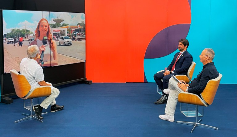 Três homens branco sentados, um deles vestindo um blazer azul e outros blusa socia. Ao fundo uma tela com uma mulher ao microfone.