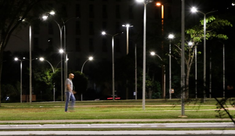 Na foto um homem caminha a noite sob as luzes de um parque