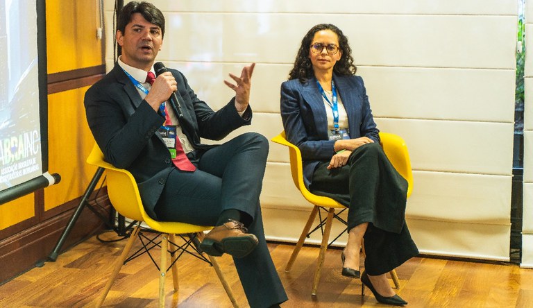 Duas pessoas sentadas, uma delas segura um microfone para falar o público do evento Summit Abrainc.