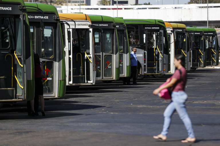 A imagem mostra um ônibus em destaque, com uma mulher ao fundo caminhando e o motorista visível na frente.