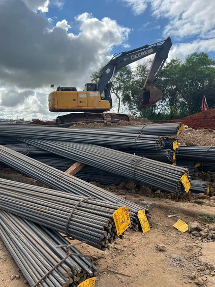 Na foto vergalhões de aço em primeiro plano com trator ao fundo trabalhando