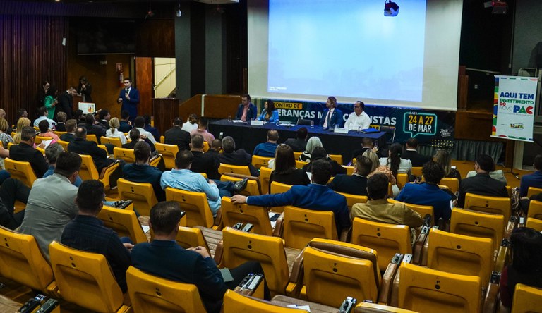 A imagem mostra um evento em um auditório com quatro palestrantes sentados em uma mesa e um apresentador falando ao público. Ao fundo, há uma tela de projeção e banners indicando que o evento é sobre o "Novo PAC" e um "Encontro de Lideranças".
