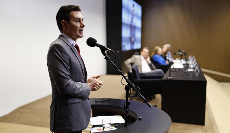 Homem branco vestindo um blazer azul marinho falando ao microfone, no fundo quatro pessoas assistindo sentados numa mesa.