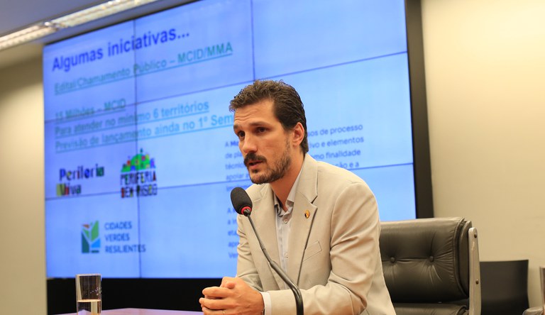 Homem branco sentado vestindo um blazer bege ao fundo um painel de tv.