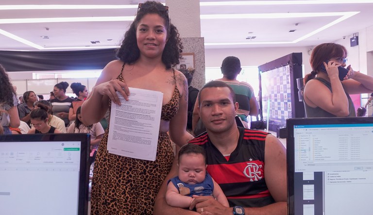 Na imagem há uma mulher negra vestindo um vestido marrom e cabelos presos, um homem sentado segurando um bebê com roupa azul.