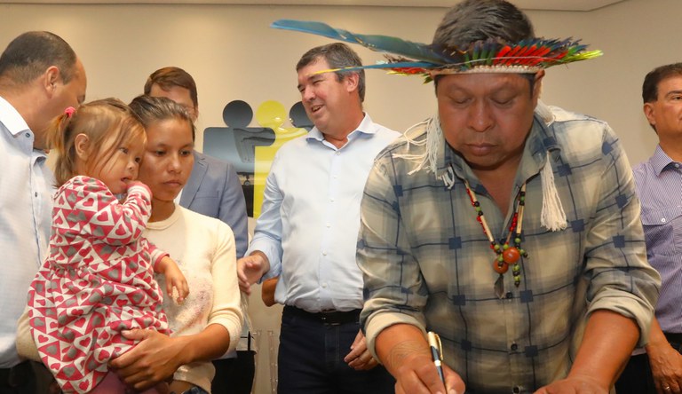 Três homens,, sendo um deles um cacique com cocá assinando um documento. Ao lado uma mulher com vestido rosa segurando sua filha no colo.