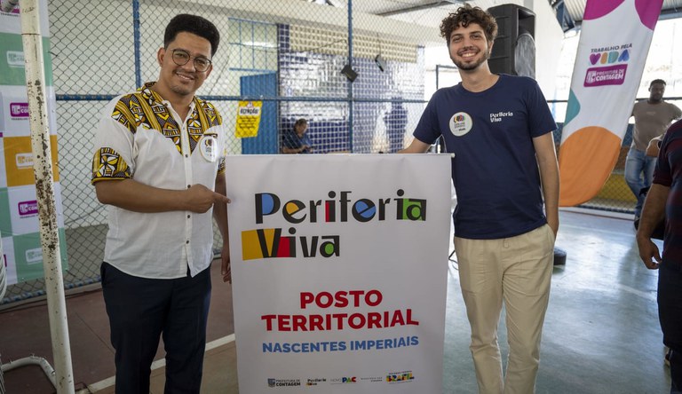 Um homem negro vestido com uma blusa branca com detalhes vermelho ao lado de um homem branco com uma blusa azul. No meio um banner com a palavra Periferia Viva.