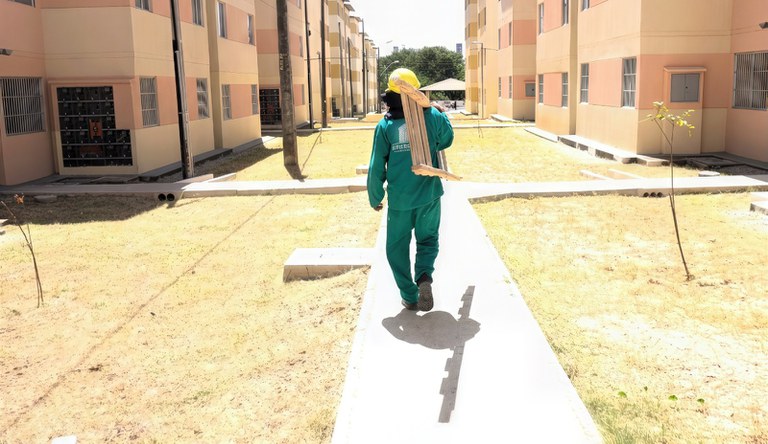 Um homem com macacão verde carregando uma escada no ombro andando rumo ao conjunto habitacional.