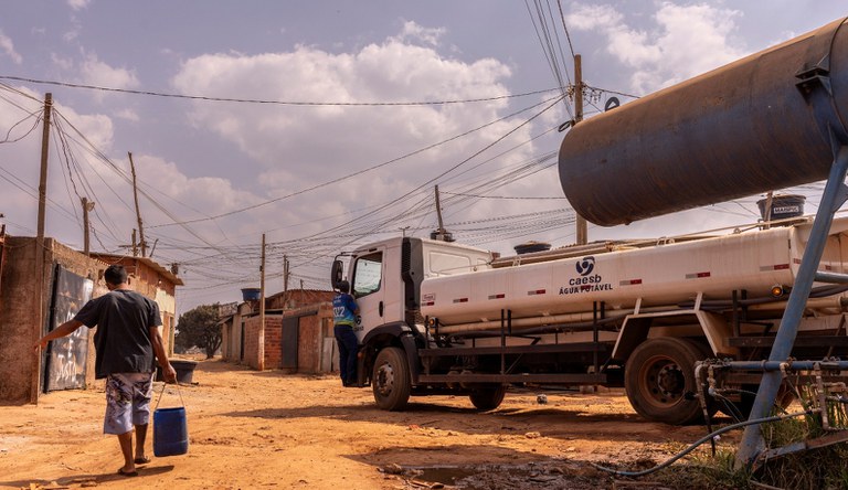 Homem vestindo uma blusa e um short carregando um balde de água. Ao fundo um caminhão pipa com símbolo da CAESB.
